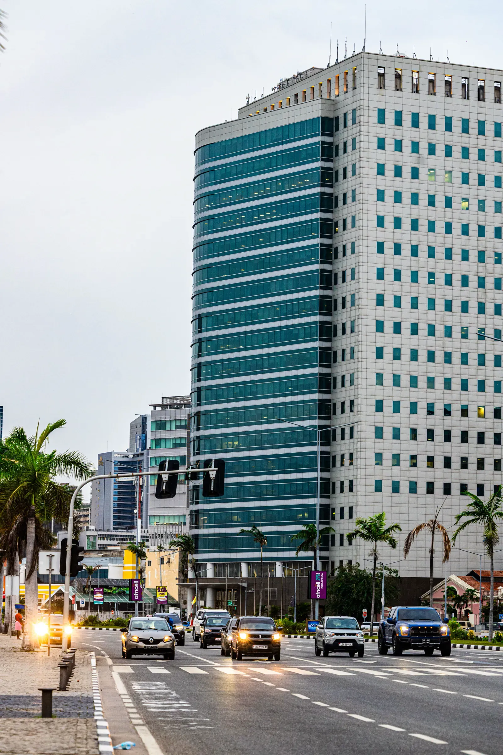 Avenida moderna de Luanda com torres de escritórios, palmeiras e trânsito.